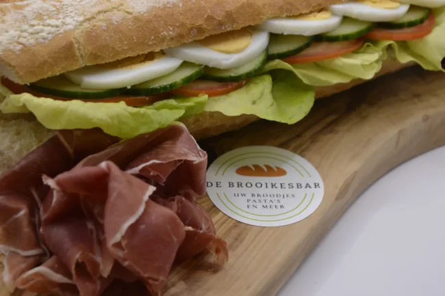 Broodje met sla, tomaat, komkommer en ei, met prosciutto op een houten plank.